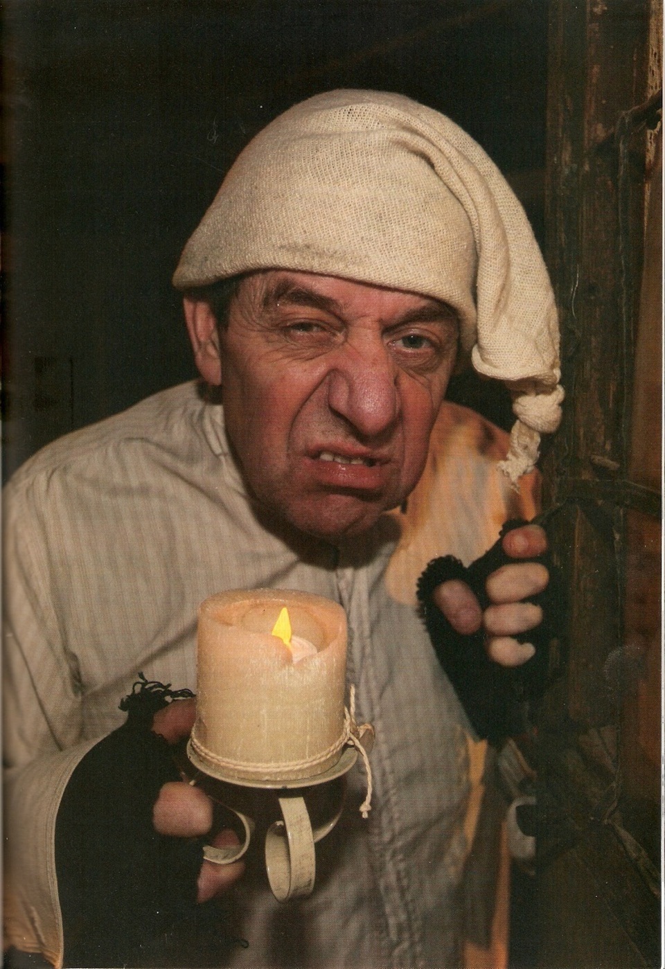 Person dressed in old-fashioned nightwear and fingerless gloves, wearing a nightcap and holding a lit candle in a holder, peering forward with a curious or suspicious expression.